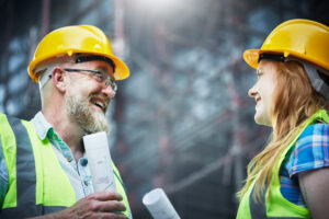 A mature male construction worker and a young female colleague stand by scaffolding on an unfinished building looking at each other and smiling in satisfaction.